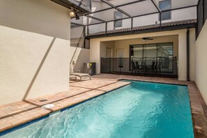 Una piscina con deck de azulejos, un patio cubierto y un cerramiento de vidrio que da a un espacio interior con ventilador de techo.