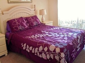Una cama con colcha de flores moradas, cabecera blanca y lámpara de noche.
