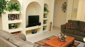 A living room with a sofa, a coffee table with flowers, a TV, and shelves with plants and books.