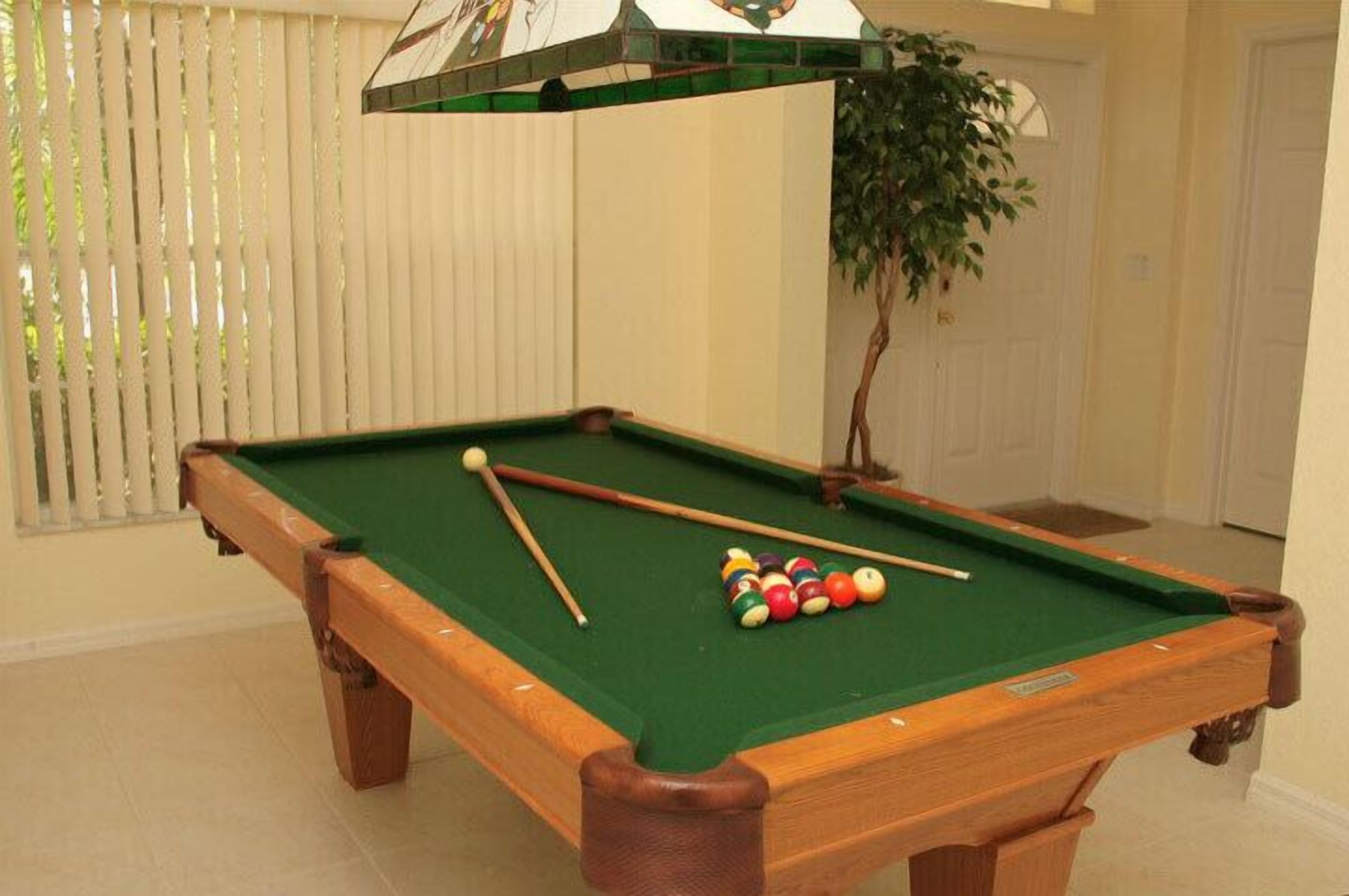 A pool table with balls and cues, a potted plant, and a window with blinds.