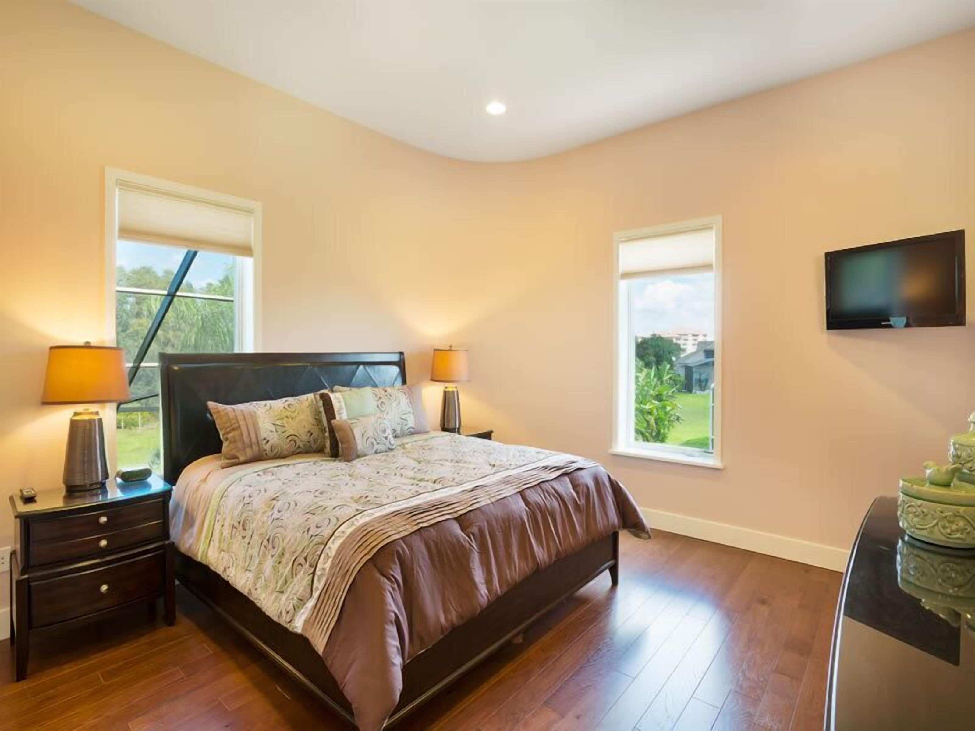 A bedroom with a large bed, two bedside tables with lamps, a flat-screen TV, and two windows offering a view of the outdoors.