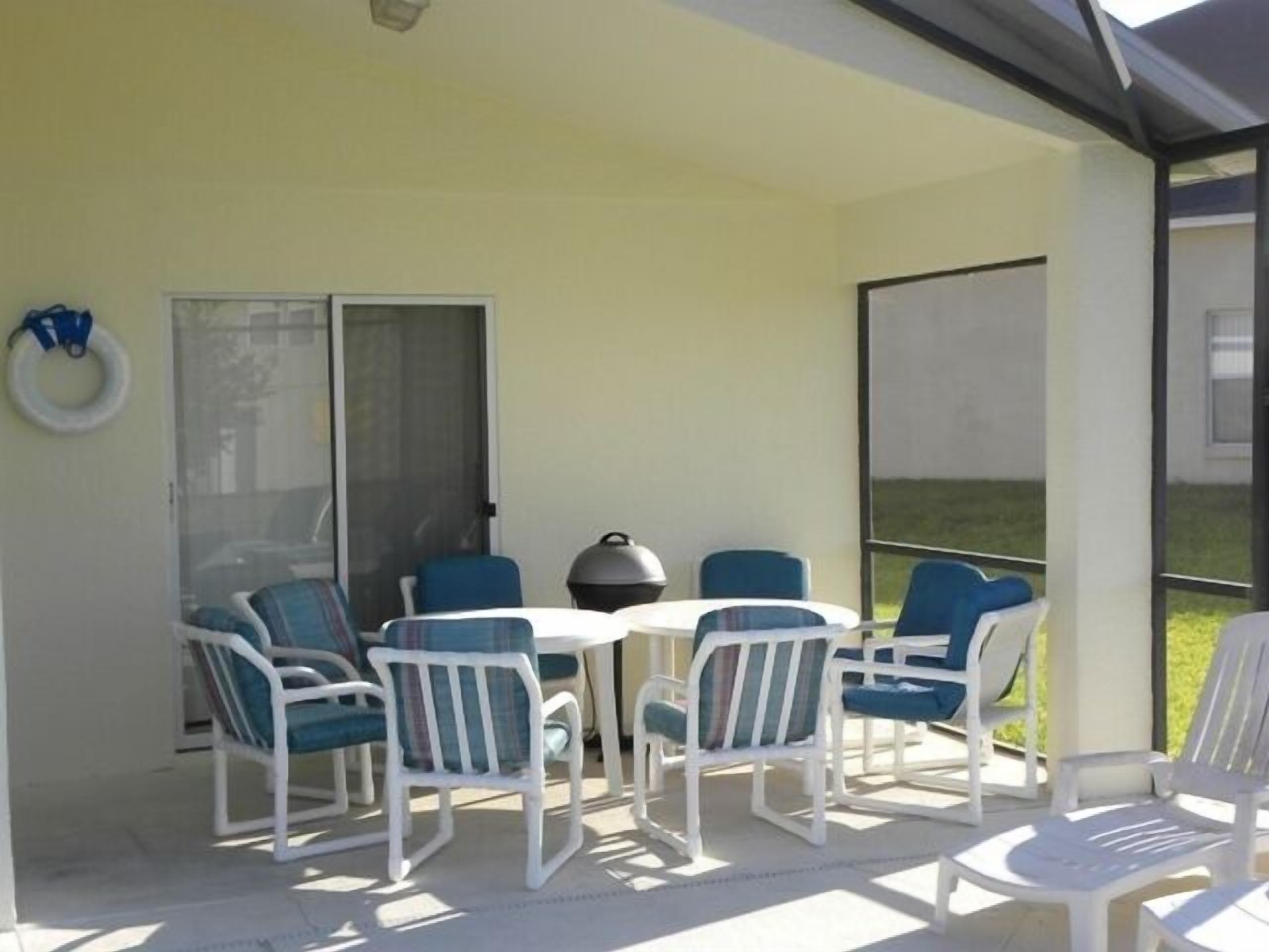 A covered outdoor seating area with white plastic chairs and a table, a grill, and a view of a grassy area.