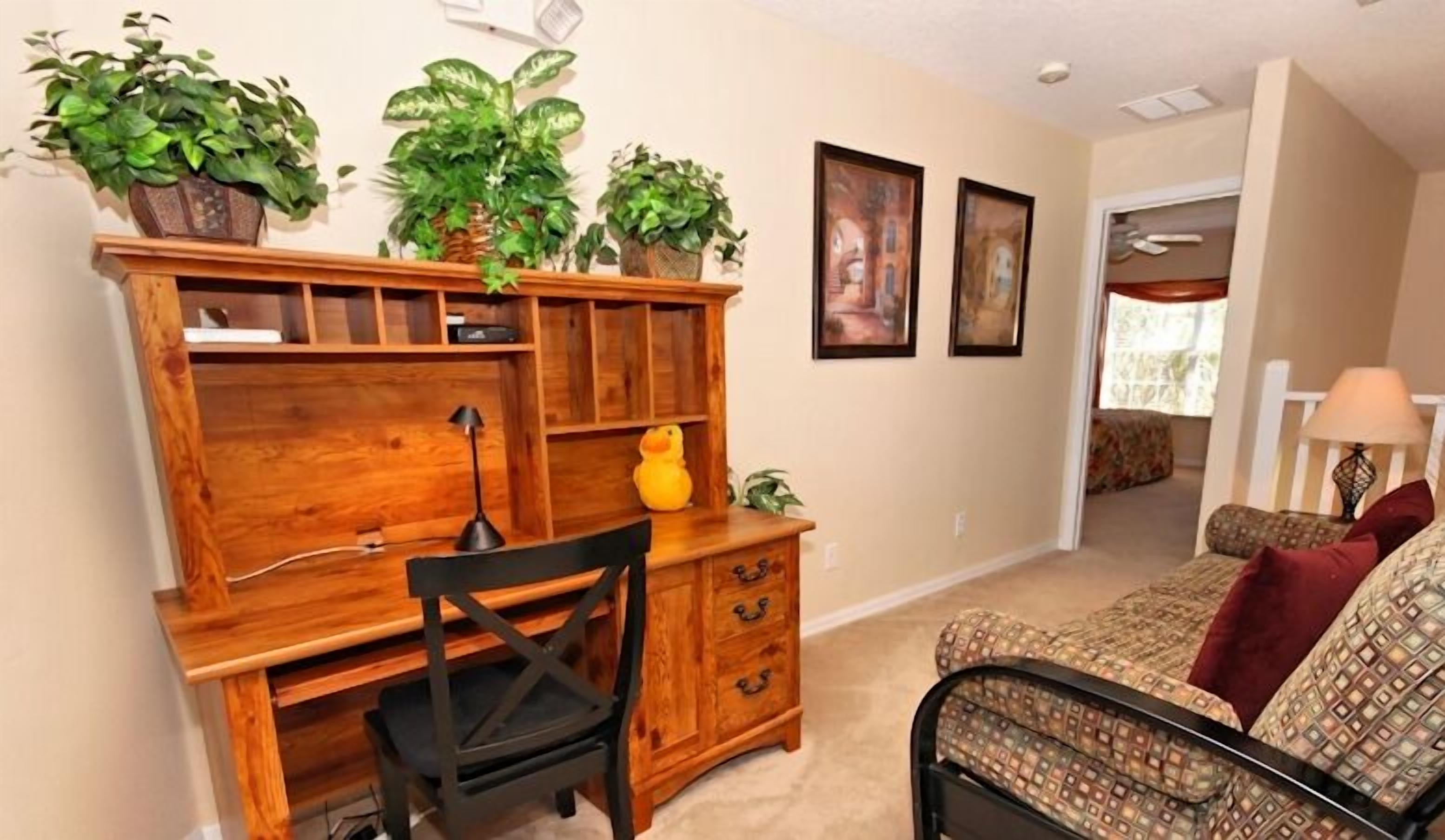 A room with a wooden desk, a chair, a sofa, and a lamp. There are several potted plants on the desk and a painting on the wall.