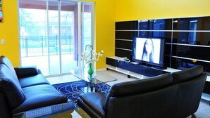 Una sala de estar con un televisor de pantalla plana grande, sofás de cuero negro, una mesa de centro de vidrio y un gran ventanal corredizo que da a un balcón.