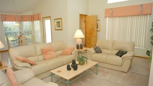 A living room with two beige sofas, a coffee table, and a lamp. There is a door leading to another room, and a window with blinds. - Ov64 - Oak Island Harbour - 3 Bed 2.5 Baths Villa (Kissimmee)