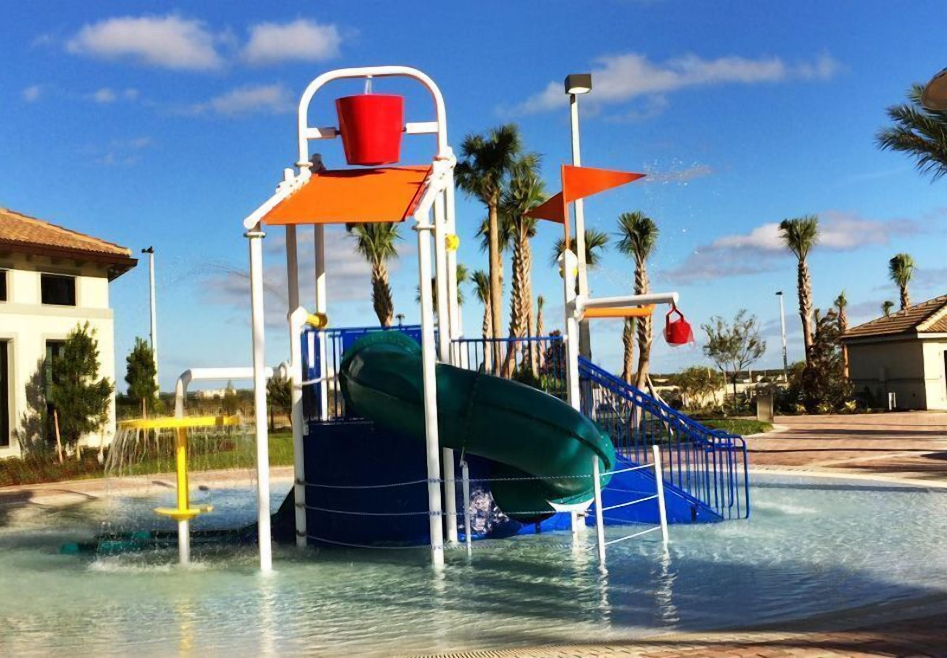 A water play area with a slide and splash pad, surrounded by palm trees and buildings.