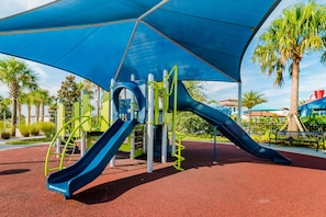 Un terrain de jeux avec un toboggan bleu et des structures d’escalade, entouré de palmiers et bordé d’un pavé rouge.