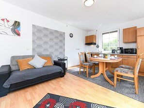 A modern living room with a sofa, a dining table with chairs, a kitchen area with cabinets and appliances, and a large window providing natural light.