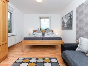A bedroom with a wooden bed, a gray sofa, a patterned rug, and a window with blinds.