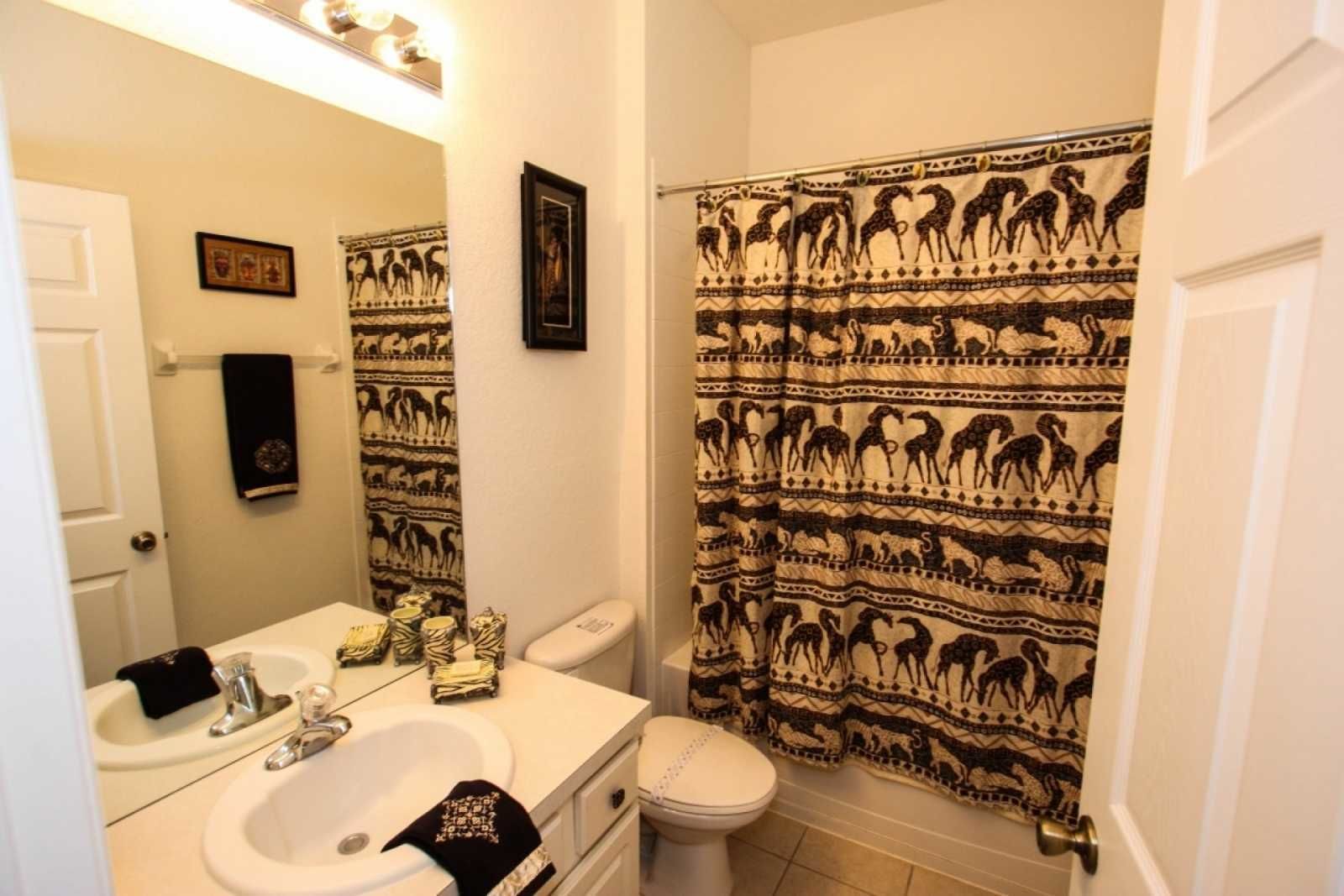Un baño con lavamanos blanco, espejo e inodoro. Se aprecia un perchero con una toalla negra y un cuadro enmarcado en la pared. La cortina de ducha tiene un diseño estampado.
