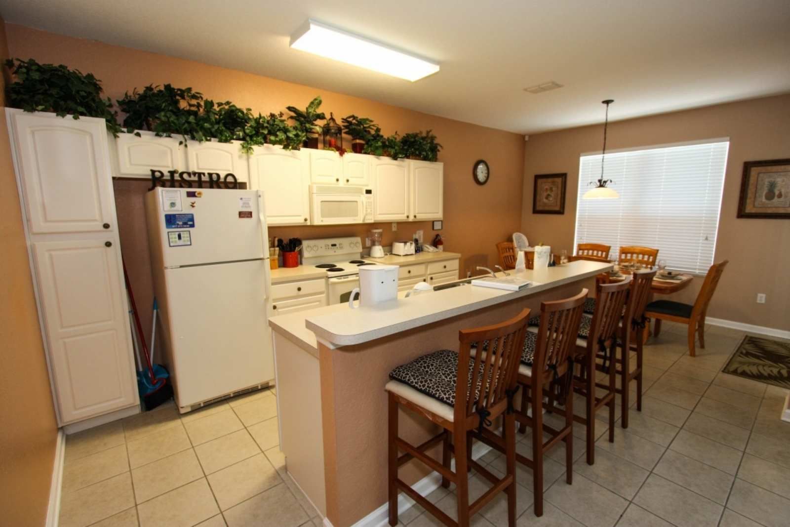 Una cocina con armarios blancos, un refrigerador, un microondas y una estufa. Hay una zona de comedor con sillas de madera y una mesa.