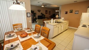 A dining area with a wooden table set for four, a kitchen with white cabinets, and a living area with a sofa and a TV.