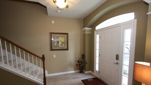 A hallway with a wooden staircase, a framed picture on the wall, a door with a window, and a lamp on the right.