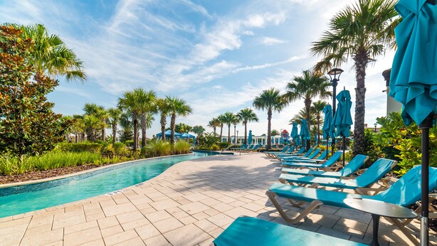 A poolside area with lounge chairs and umbrellas, surrounded by palm trees and a clear sky.