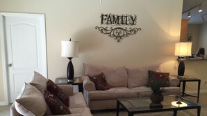 A living room with a beige sofa, a matching armchair, a coffee table, and two side tables with lamps. A decorative wall piece with the word "FAMILY" is visible.