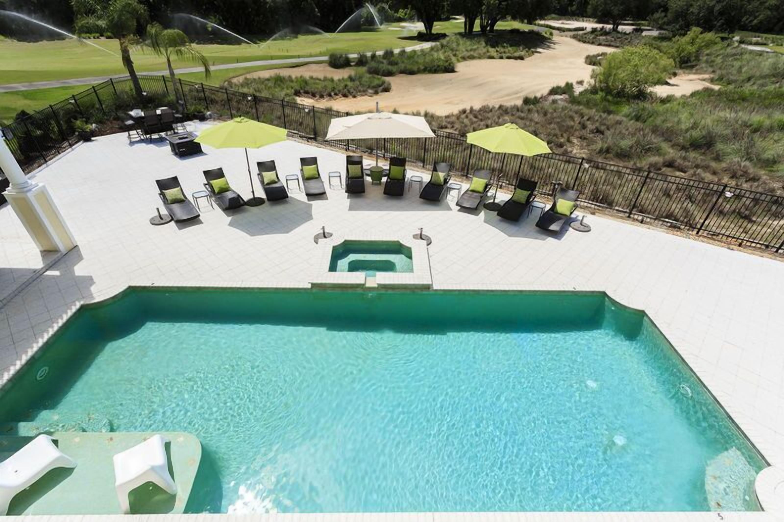 A pool area with lounge chairs and umbrellas, surrounded by a fence and a golf course in the background.