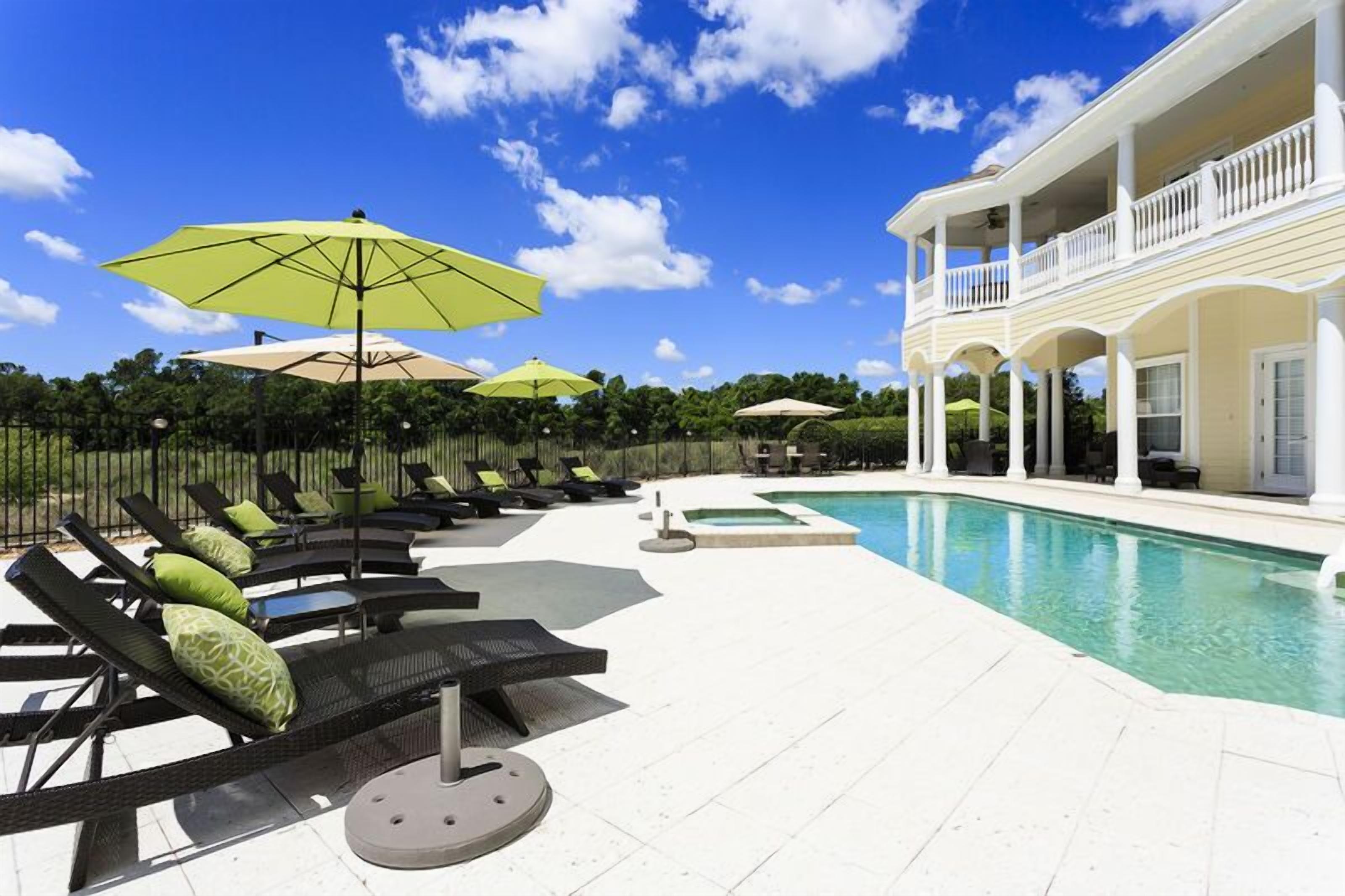 Un espace aménagé au bord de la piscine, avec des chaises longues et des parasols, une piscine, et un bâtiment doté de balcons.