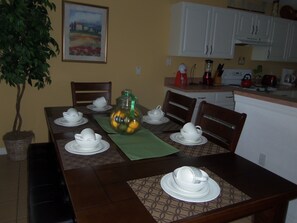 Een eettafel gedekt met witte servies, een groene placemat en een decoratieve glazen kruik met fruit.