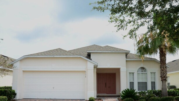 A single-story house with a white garage door, a brown front door, and a small front yard with a palm tree and shrubs. - Ov3786 - Cumbrian Lakes - 4 Bed 3.5 Baths Villa (Kissimmee)