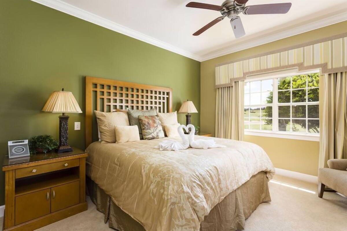 A bedroom with a large bed, two bedside lamps, a wooden headboard, a ceiling fan, and a window with curtains.