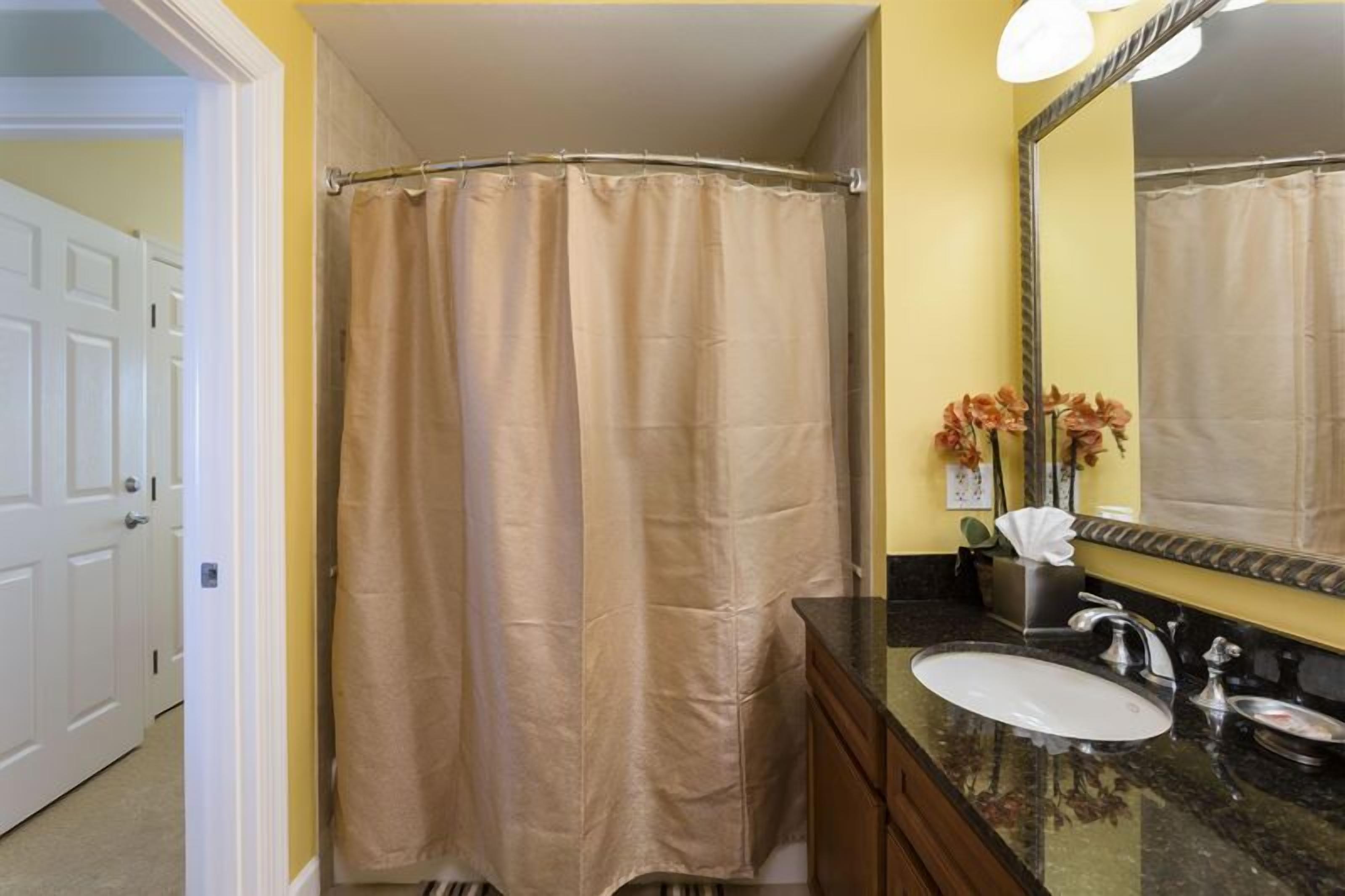 Une salle de bain avec un rideau de douche beige, un lavabo blanc, un miroir et un vase avec des fleurs.