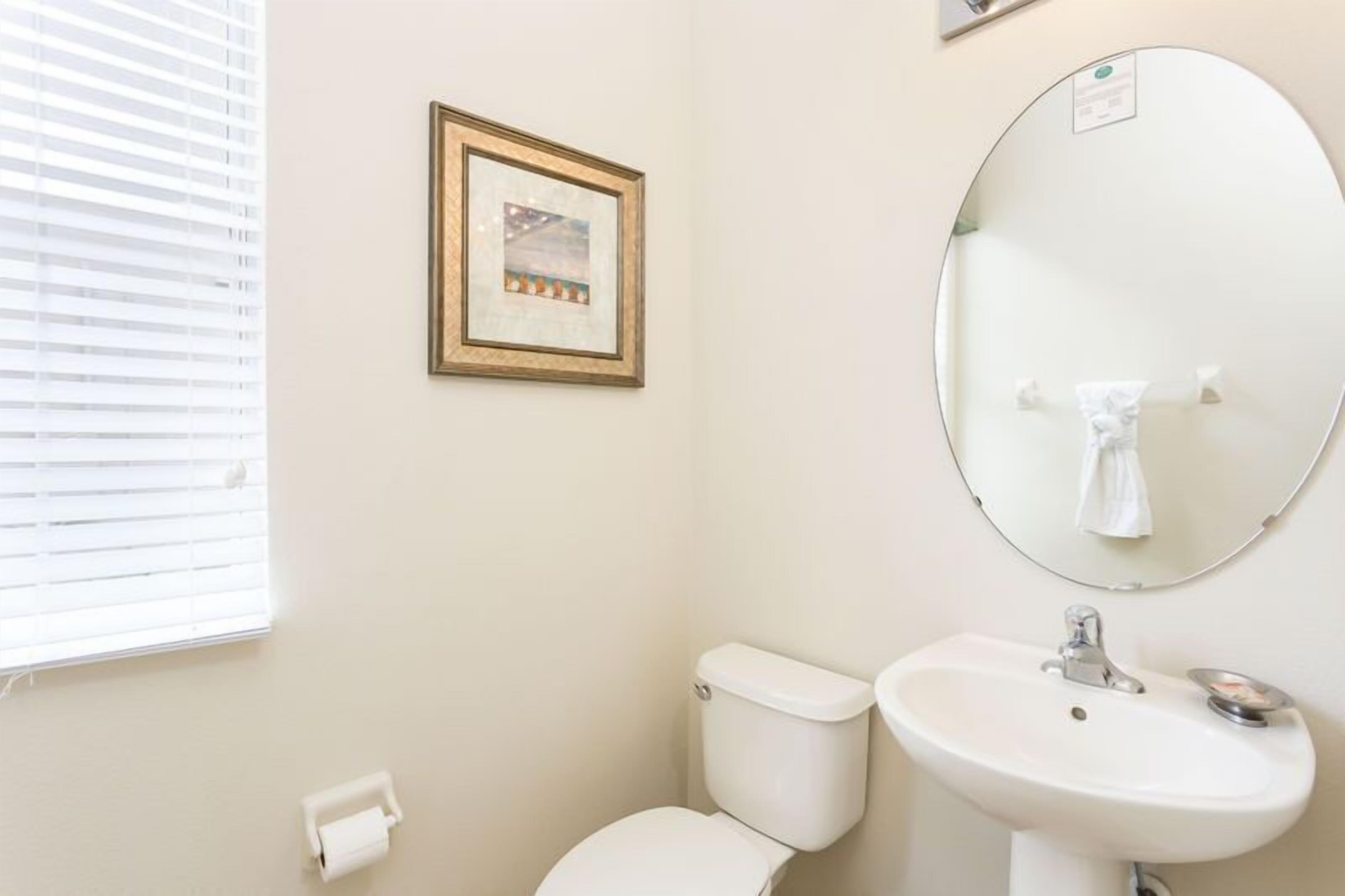 Une salle de bain avec des toilettes, un lavabo et un miroir. Un tableau encadré est accroché au mur, et il y a une fenêtre avec des stores.