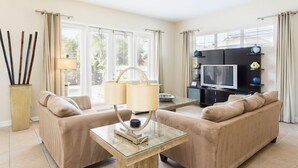Ein modernes Wohnzimmer mit beigen Sofas, einem Glastisch, einem Fernseher und einem großen Fenster mit Vorhängen.