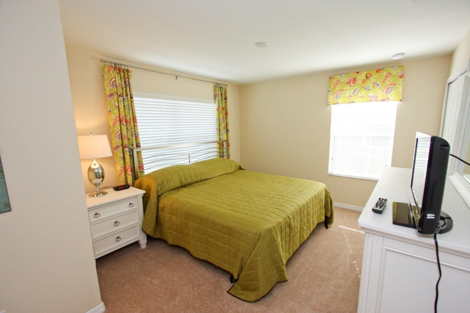 A bedroom with a bed, a bedside table with a lamp, a dresser, a TV, and a window with curtains.