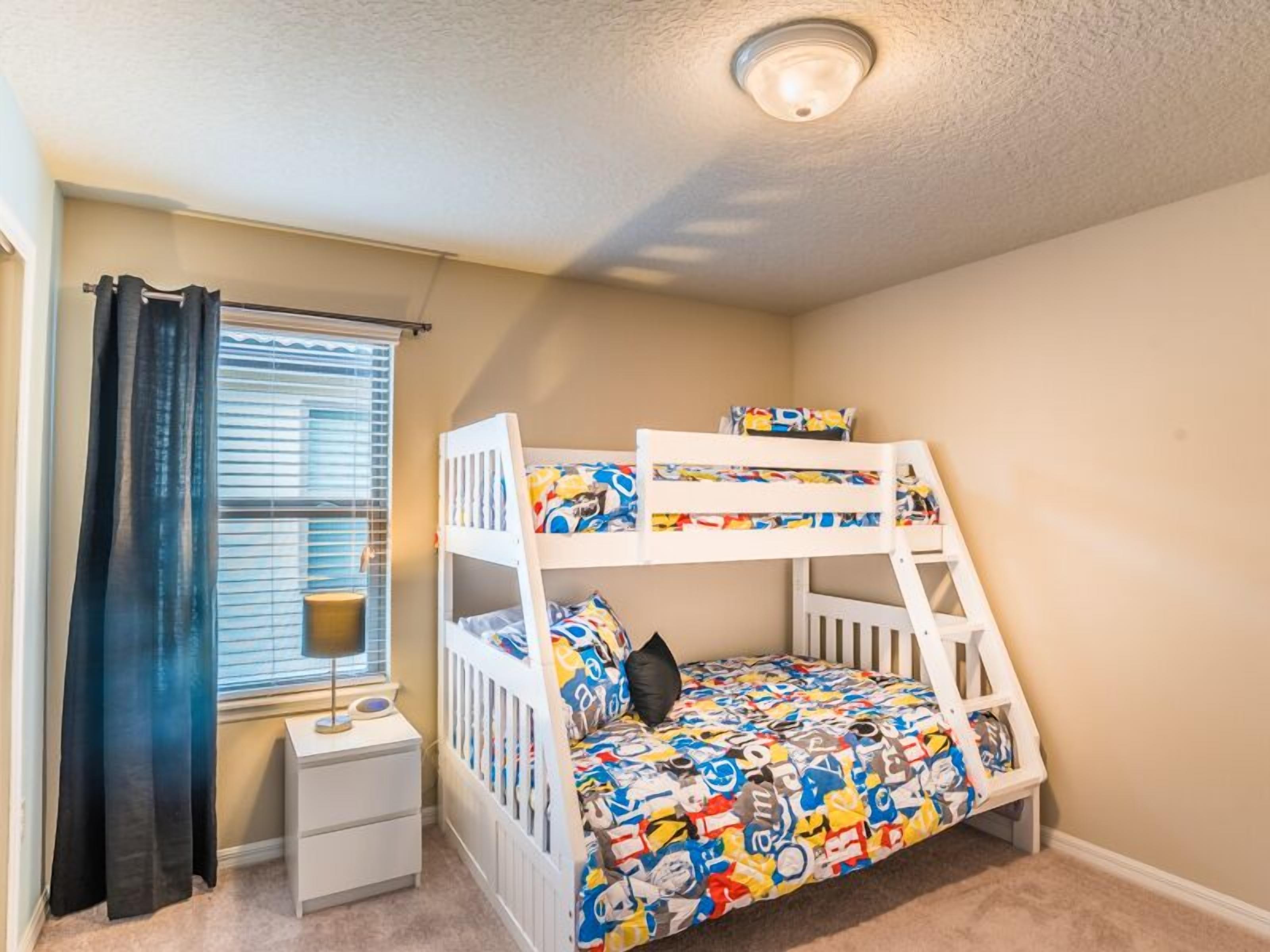 A bunk bed with a colorful mattress, a bedside table, and a window with blinds.