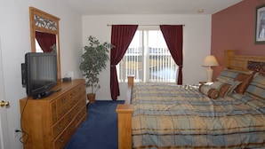 Une chambre à coucher avec un lit, une commode, un téléviseur, une plante et une fenêtre avec des rideaux.