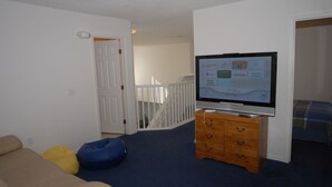 Une pièce avec un téléviseur posé sur une commode en bois, un canapé beige et une moquette bleue.