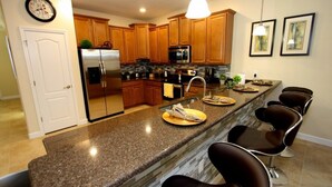 A modern kitchen with granite countertops, wooden cabinets, a stainless steel refrigerator, and a built-in microwave. There are four bar stools and a clock on the wall.