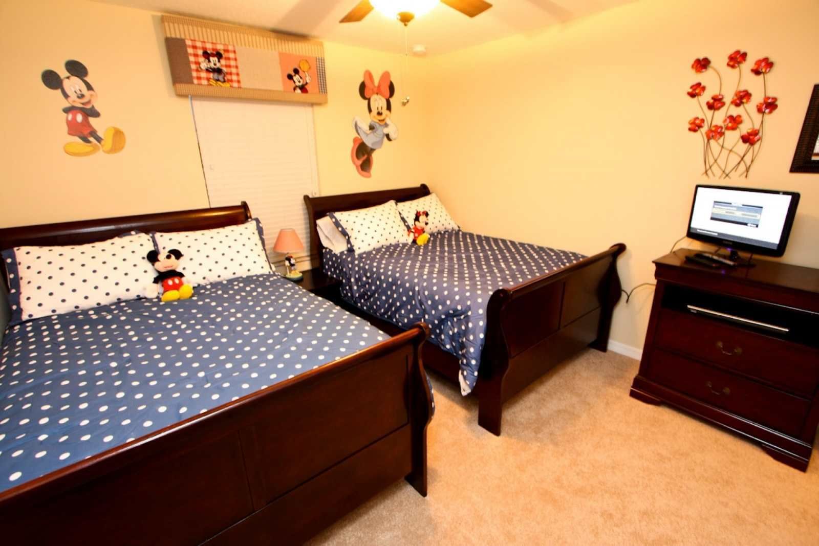 Een slaapkamer met twee houten bedden, elk voorzien van een Mickey Mouse pluche speeltje. Er is een televisie op een dressoir en een muur met decoraties van Mickey Mouse en Minnie Mouse.