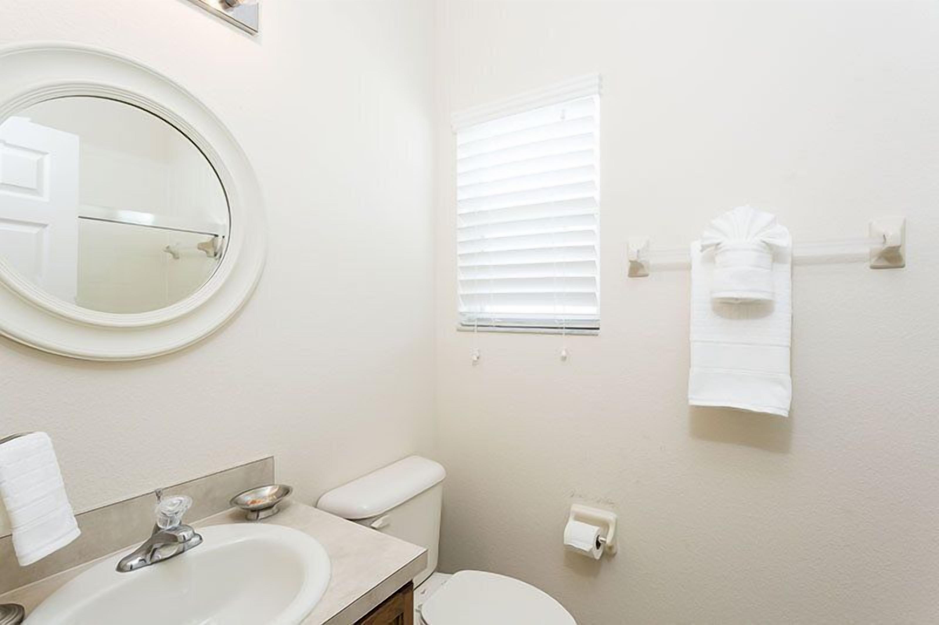 Un baño con un espejo redondo, un lavabo, un inodoro, una ventana con persianas y un toallero con toallas.