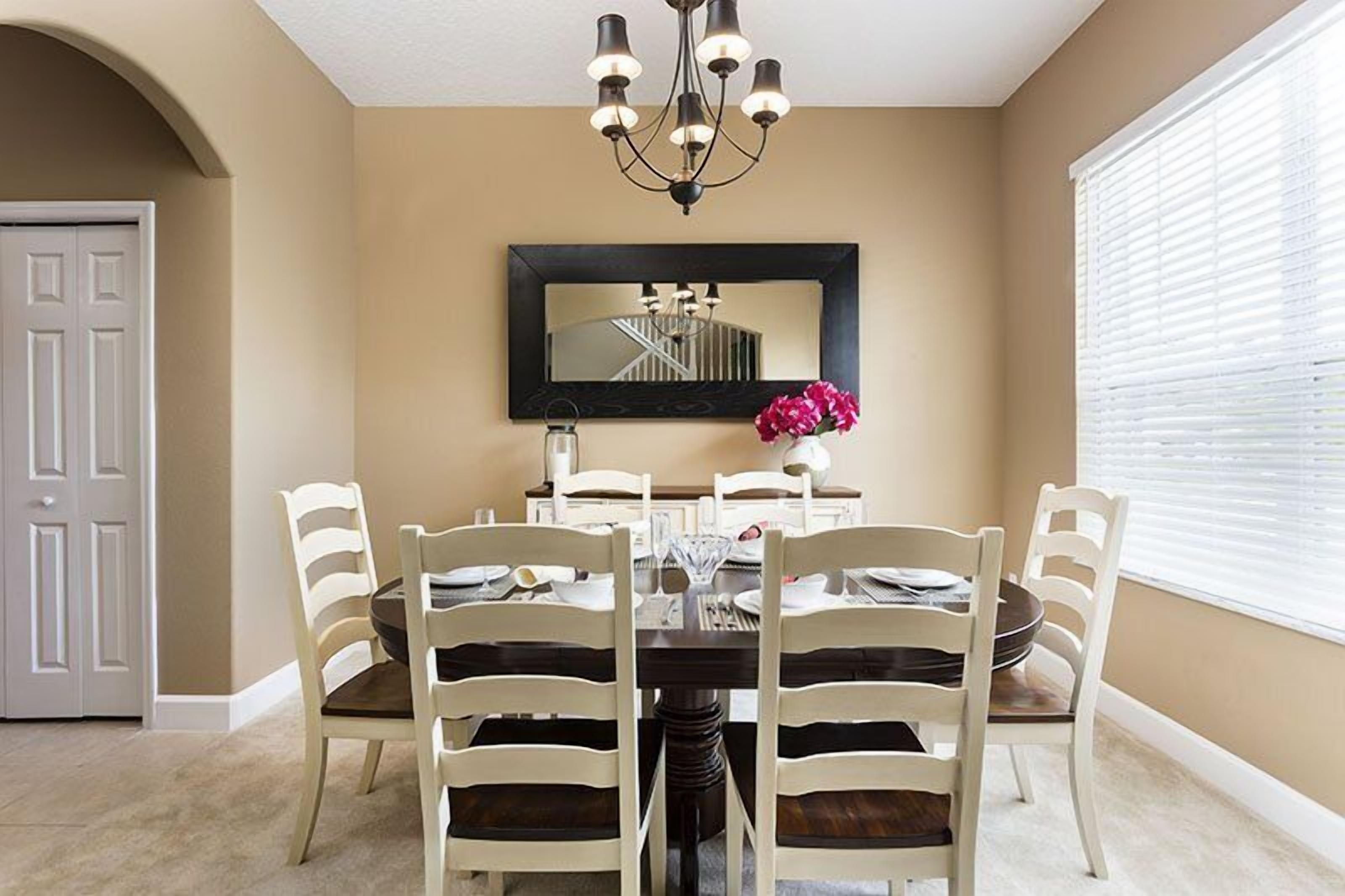 A dining room with a round table, six chairs, a chandelier, a mirror, and a vase with flowers.