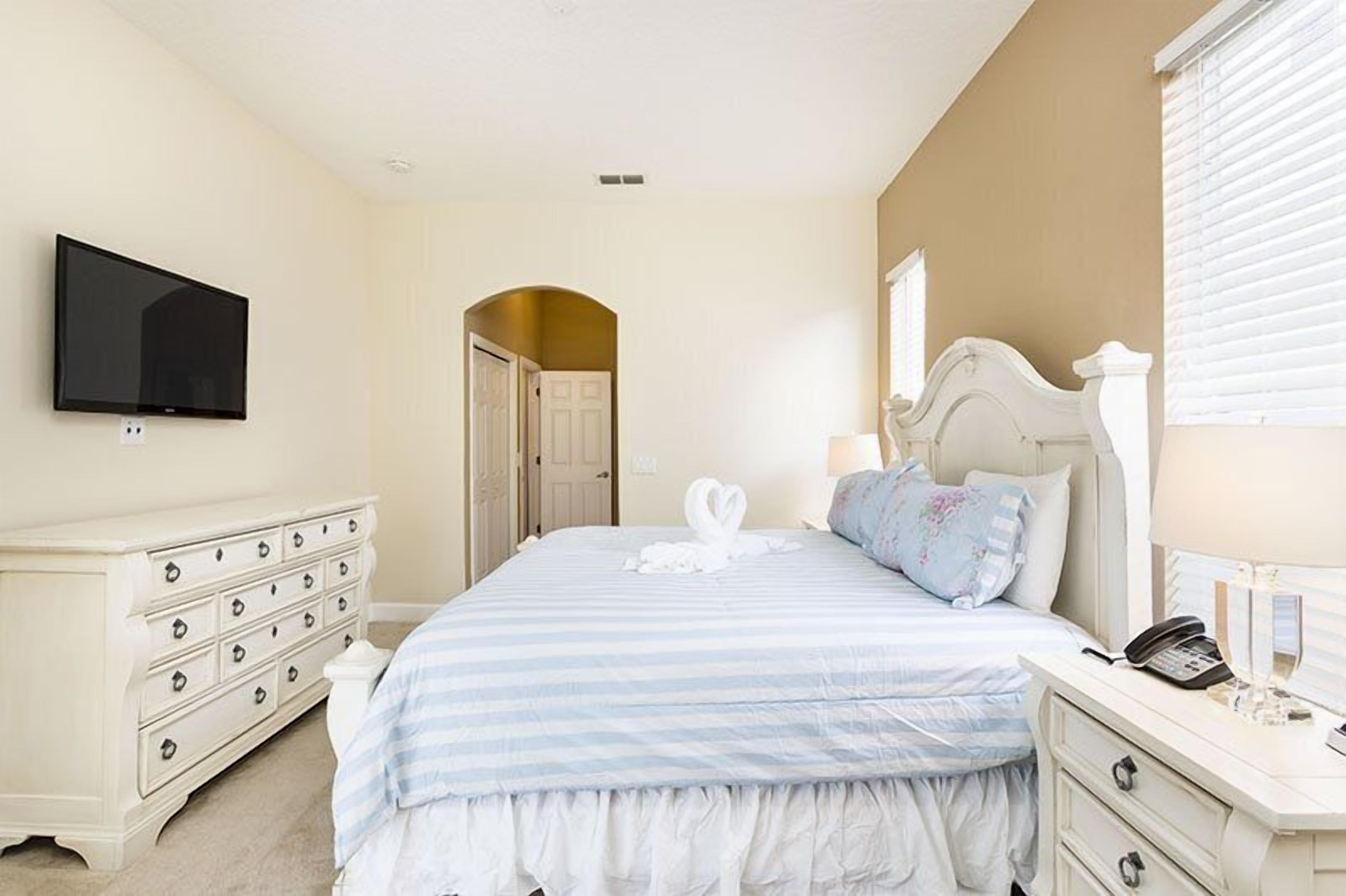 A bedroom with a bed, a dresser, a TV, and a door leading to another room.