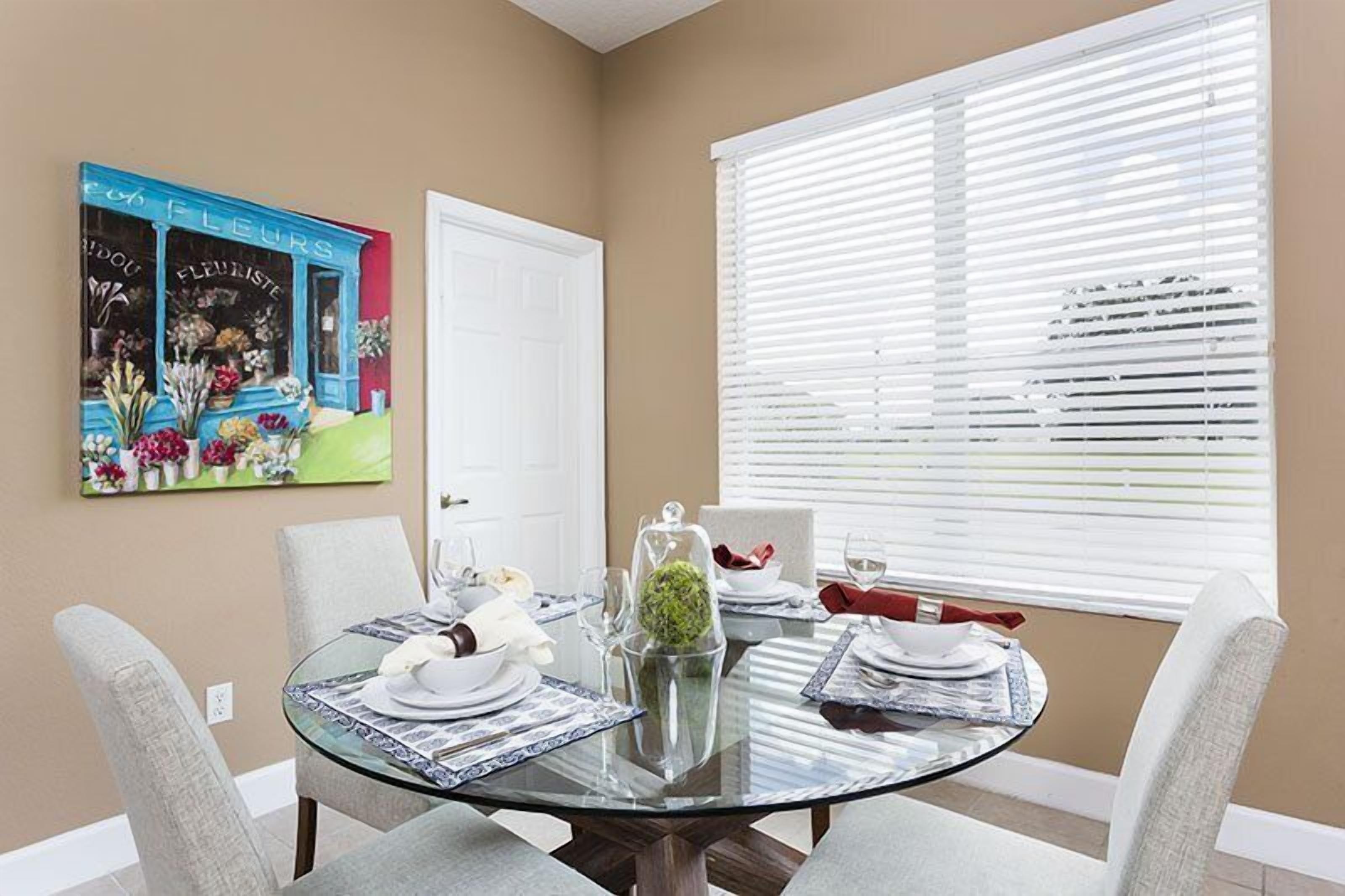 A dining area with a glass table, white chairs, a painting of flowers, and a window with blinds.