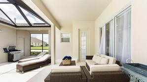 Een zonnige kamer met een bank, rieten stoelen, een barbecue en uitzicht op een huis.