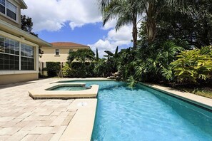 A swimming pool with a hot tub, surrounded by lush greenery and a house in the background.