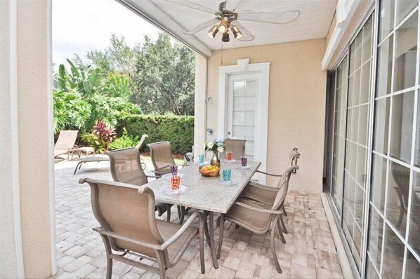 An outdoor patio with a table and chairs, a ceiling fan, and a view of greenery.