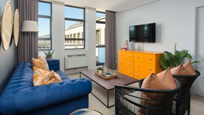A modern living room with a blue sofa, a wooden coffee table, a black wicker chair, and a bright orange cabinet. - Fountain Suites 1001 (Cape Town)