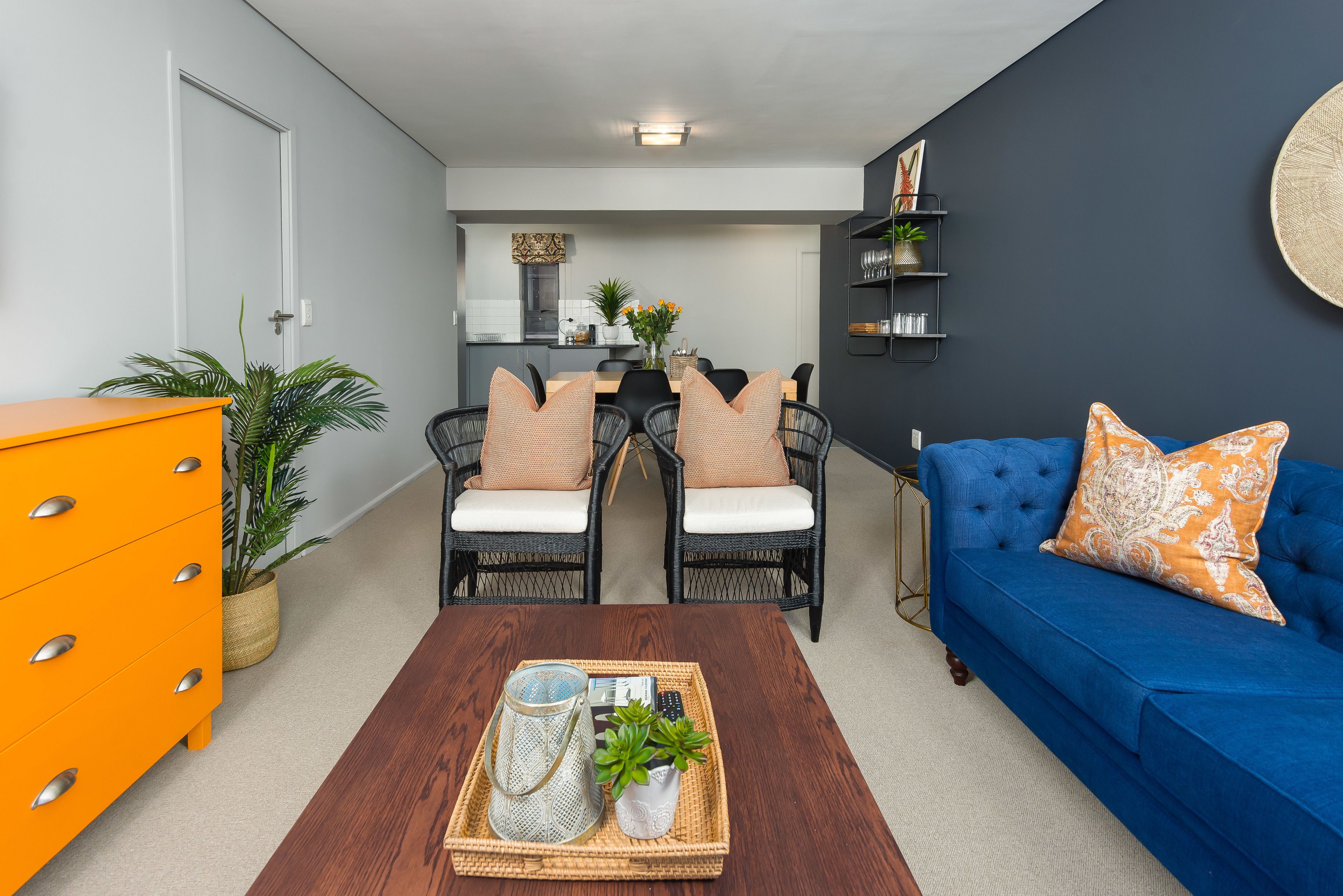Un salon avec un canapé bleu, une commode orange et un coin repas comprenant des chaises et une table en osier.