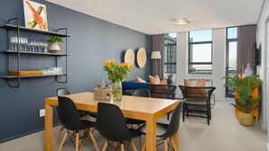 A modern living room with a dining area, featuring a wooden table, black chairs, a blue sofa, and a large window offering a city view. - Fountain Suites 1001 (Cape Town)