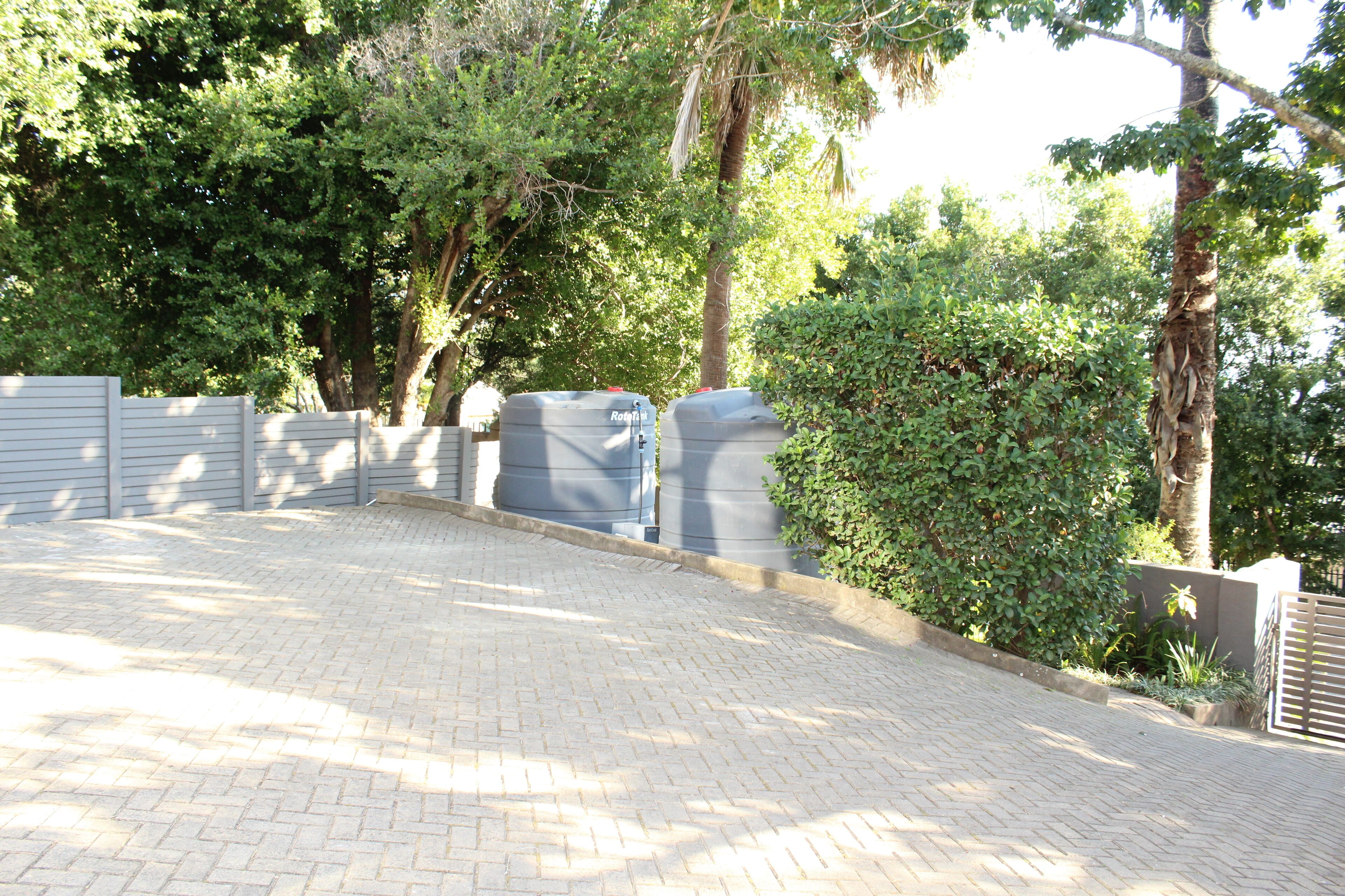 A paved driveway leading to a gated area with two large water tanks, surrounded by lush greenery and trees.