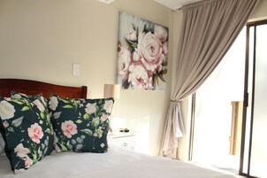 Quarto com cama, almofadas estampadas, mesa de cabeceira, abajur, pintura e janela com cortinas.