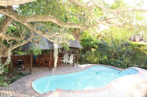Uma piscina com deck de tijolos, rodeada por vegetação exuberante e um gazebo com telhado de palha.