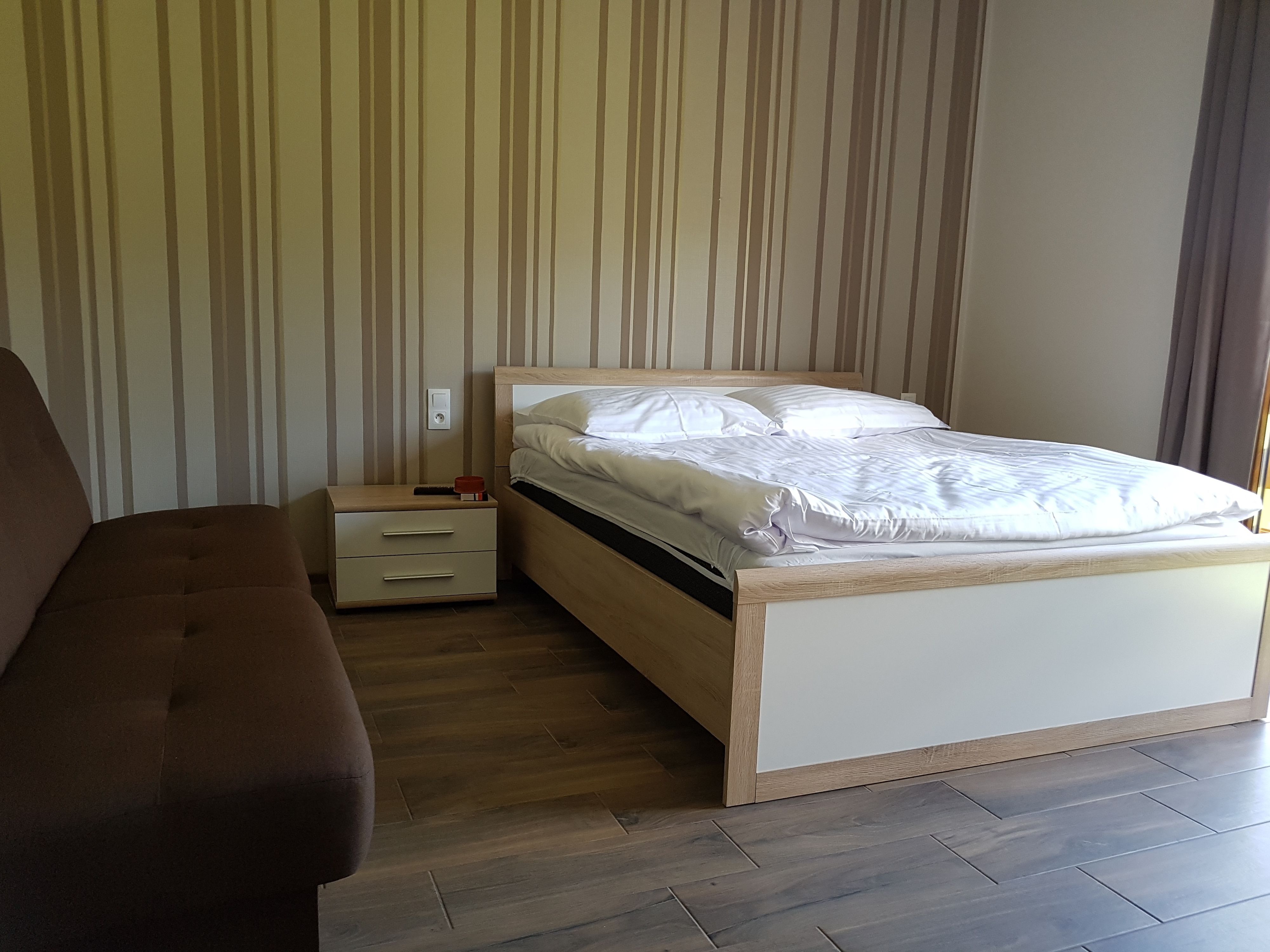 A bedroom with a bed, a nightstand, a drawer, and a brown sofa.