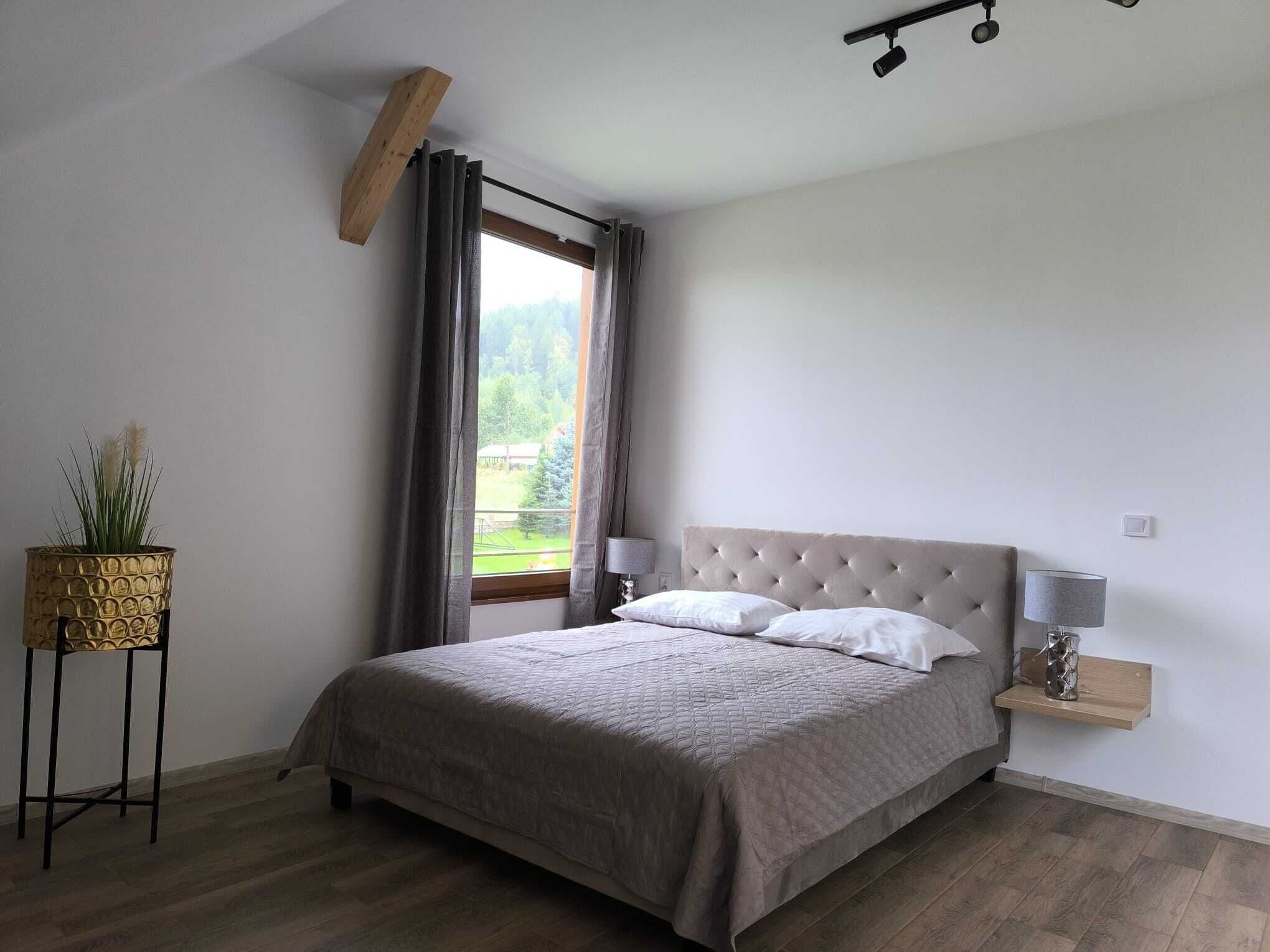 Un dormitorio con una cama grande, una mesita de noche con lámpara, una ventana con cortinas y una planta sobre un pedestal.