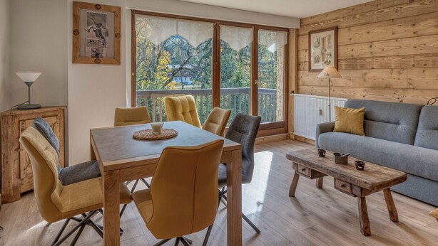 A cozy living room with a dining area, wooden walls, and large windows offering a view of trees and buildings. - 2-room apartment in Megève, south-facing balcony, near center (Megève)