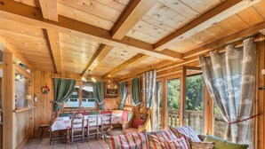 L’intérieur d’un chalet en bois chaleureux et accueillant, avec un coin repas, un canapé agrémenté de coussins à motifs et de grandes fenêtres dotées de rideaux.